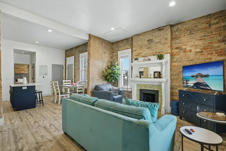 Exposed brick accent walls, wood-finish flooring, white fireplace mantel with green tile surround, recessed lighting, and a kitchen island with a white countertop