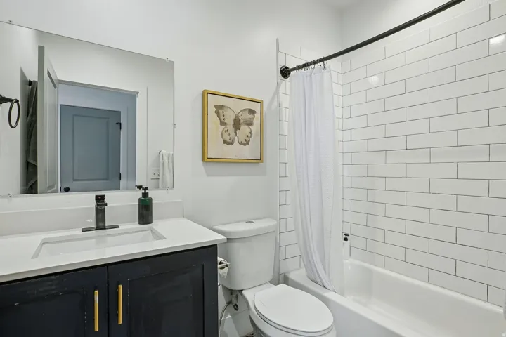 Contemporary bathroom featuring a black vanity with brass hardware and white countertop, white subway tile surround, framed mirror with integrated lighting, and a white subway tile tub/shower enclosure with chrome fixtures and white curtain rod