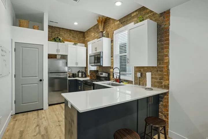 Modern kitchen featuring white cabinetry with stainless steel appliances, a white quartz waterfall-edge island with seating, wood-finish flooring, and exposed brick accent wall