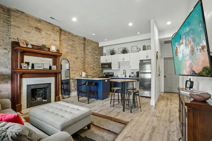 Exposed brick wall, decorative fireplace with wood mantel, wood-finish flooring, stainless steel appliances, and white cabinetry