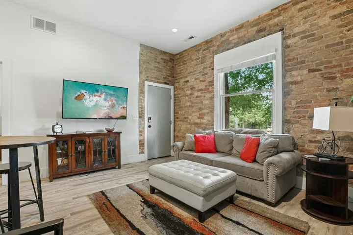 Exposed brick wall, large window with white trim, light wood-finish flooring, white painted walls, and a light gray main entry door