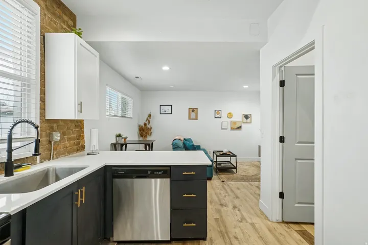 Modern kitchen featuring stainless steel appliances, black cabinetry with brass hardware, white quartz countertops, exposed brick backsplash, and wood-finish flooring throughout the open layout