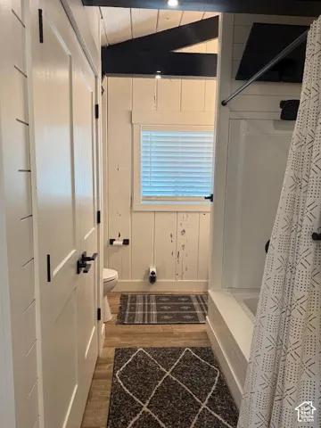 Bathroom featuring a built-in tub with a shower, wood-finish flooring, white paneling, and a window with blinds