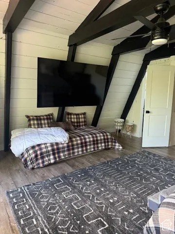 Vaulted ceiling with exposed black beams, wood-finish flooring, white shiplap walls, wall-mounted television, and a ceiling fan with integrated lighting
