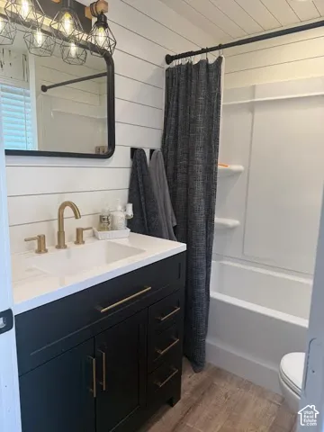 Bathroom featuring shiplap wall paneling, a dark vanity with a white countertop and gold-tone fixtures, a frameless mirror, and wood-finish flooring