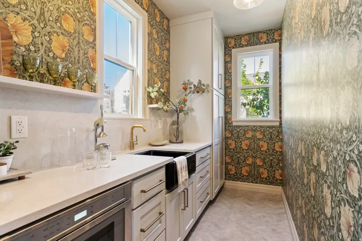 Stylish kitchen area featuring patterned wallpaper, white shaker-style cabinetry, quartz countertops, a black farmhouse sink with a gold-finish faucet, and herringbone-patterned flooring
