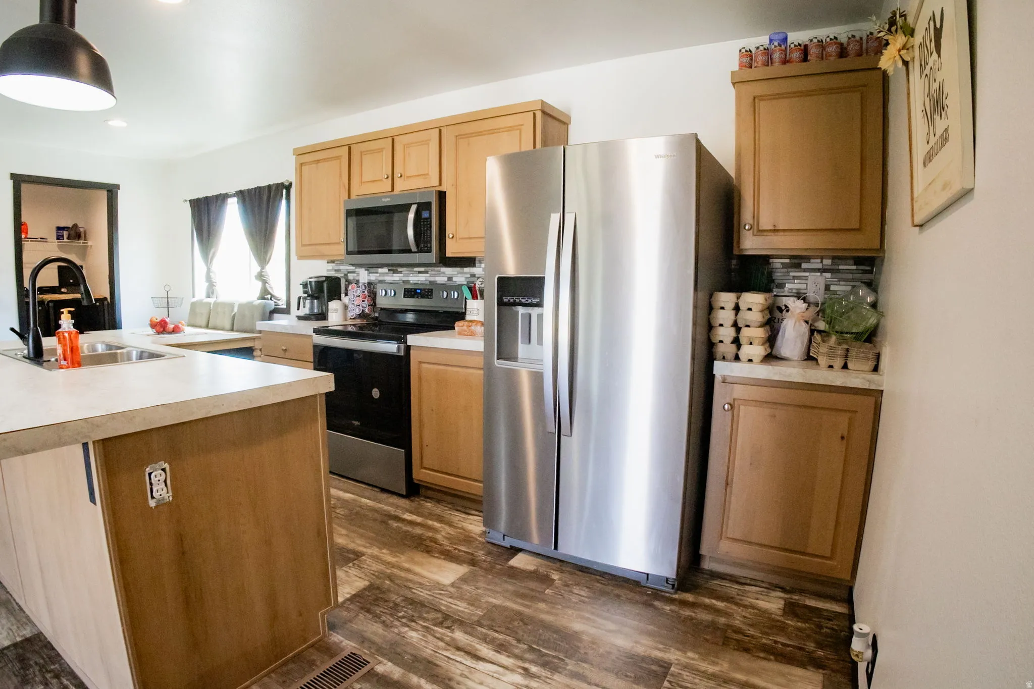 Kitchen featuring wood-finish flooring, stainless steel appliances, light wood-tone cabinetry, light-toned countertops, and a tiled backsplash