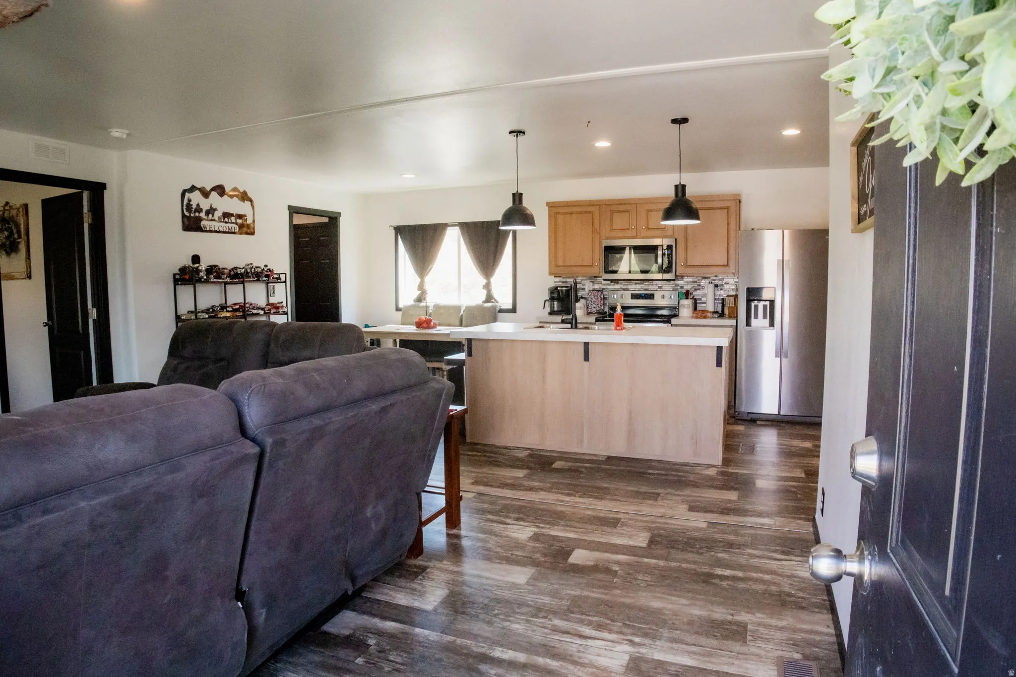 Open-concept living space featuring wood-finish flooring, a kitchen island with pendant lighting, stainless steel appliances, and light wood cabinetry