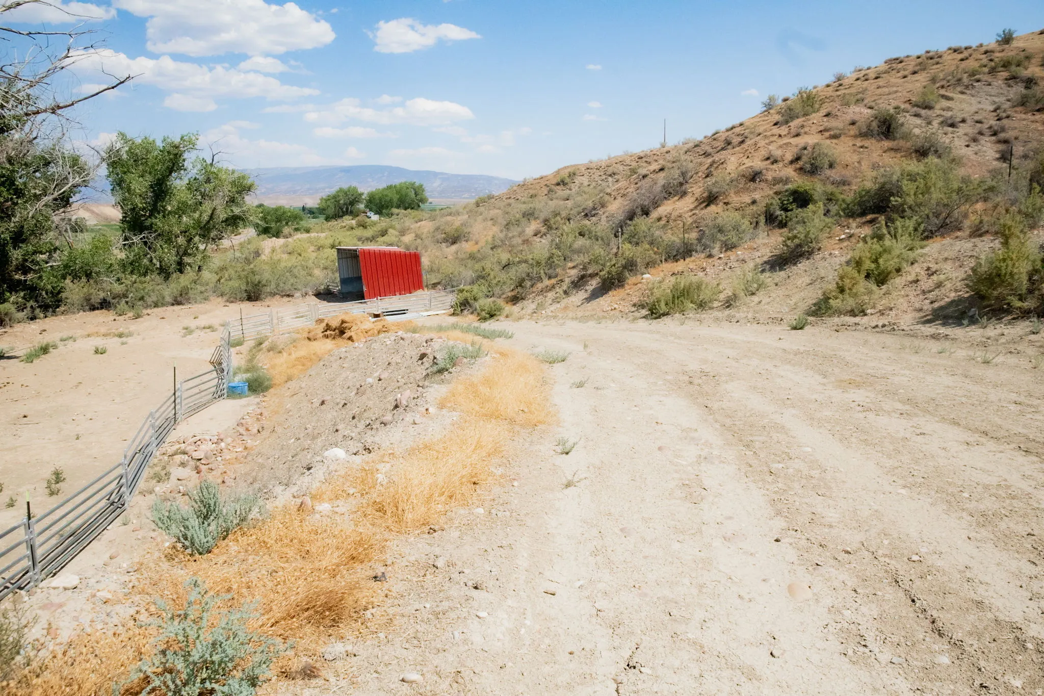Rural landscape with a dirt road, metal fencing, and a red corrugated metal structure