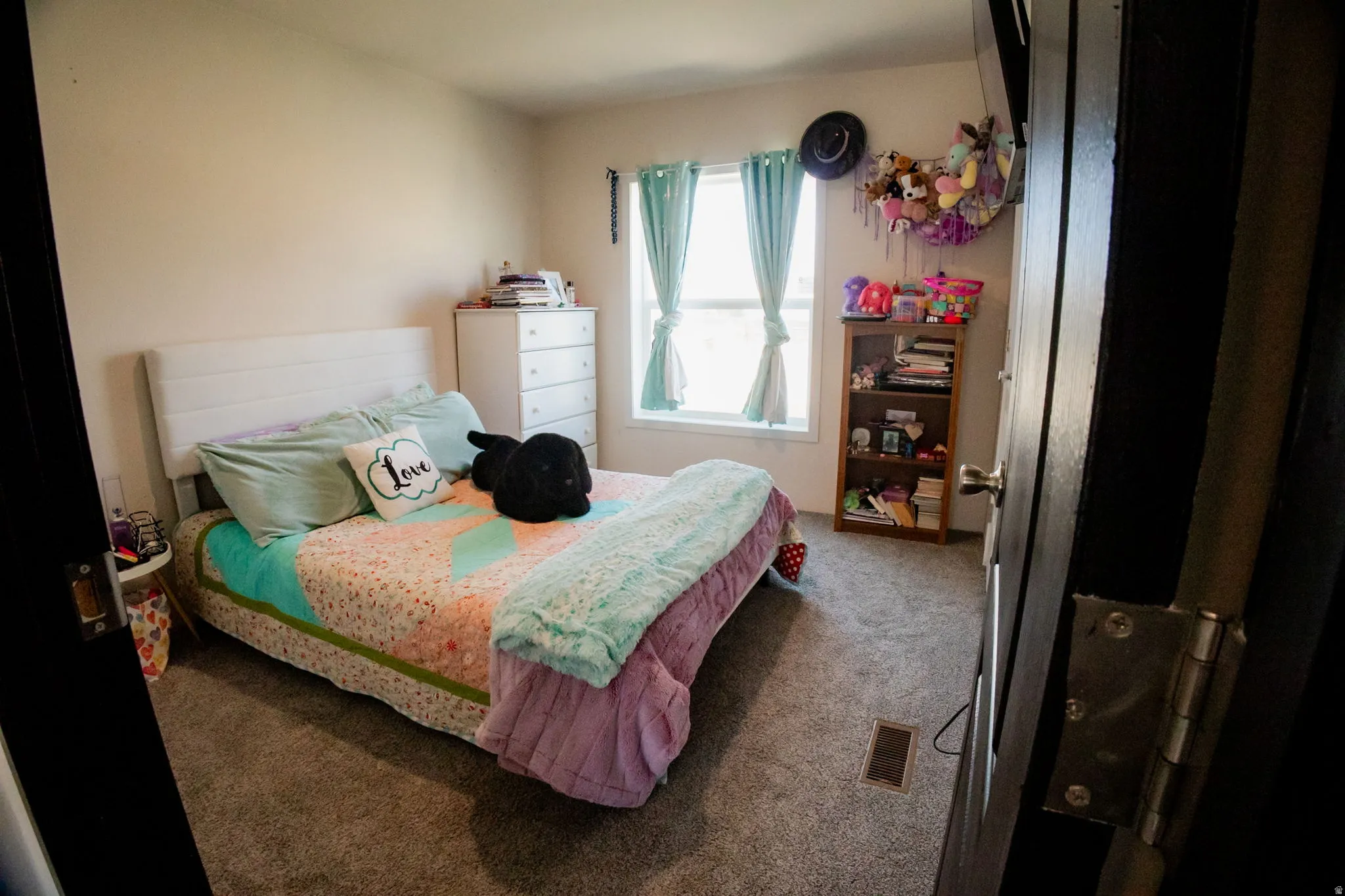 Carpeted bedroom featuring a window with light-filtering curtains, a built-in air vent, and light-colored walls