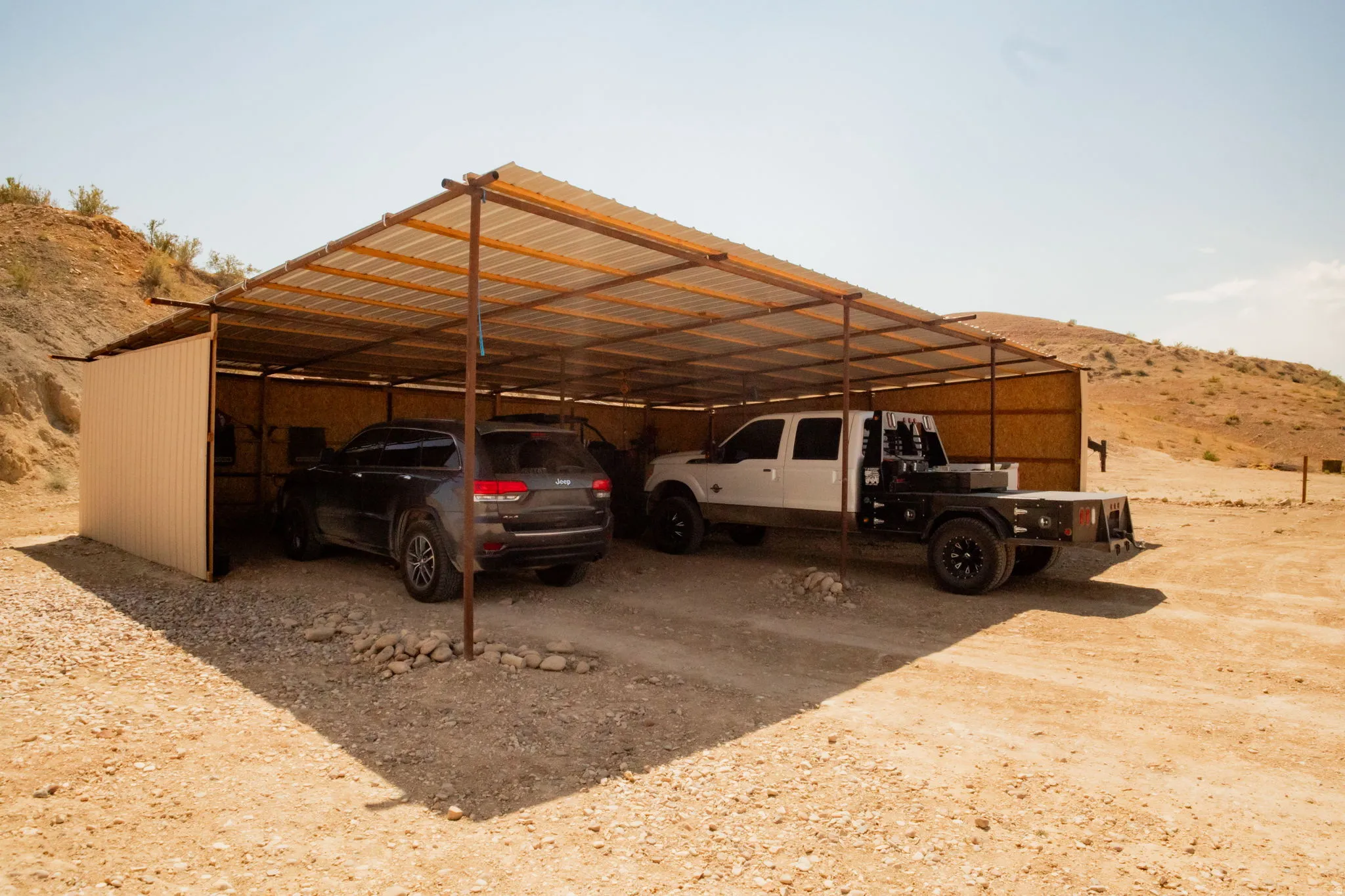 Open-air carport structure with a corrugated metal roof and a partial corrugated metal back wall, set on a gravel surface