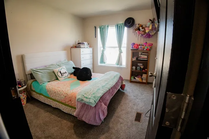 Carpeted bedroom featuring a window with light-filtering curtains, a built-in air vent, and light-colored walls