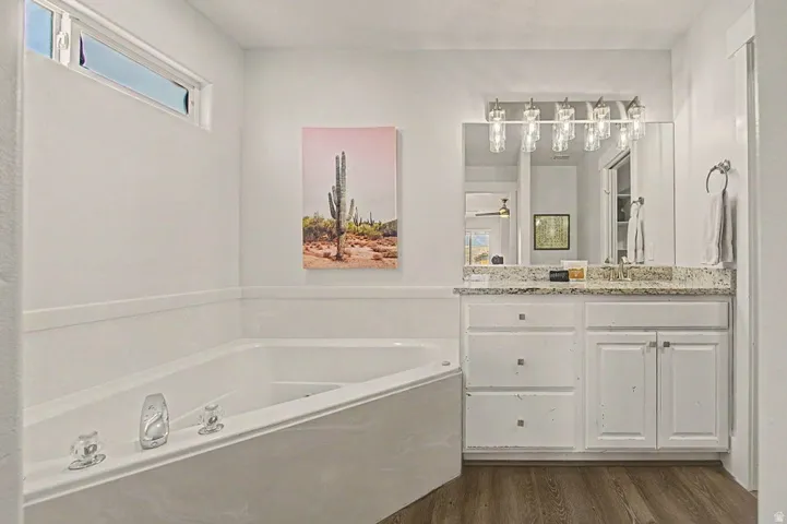 White bathroom featuring a built-in soaking tub, a vanity with a granite-style countertop, a mirror with an overhead light fixture, and wood-finish flooring