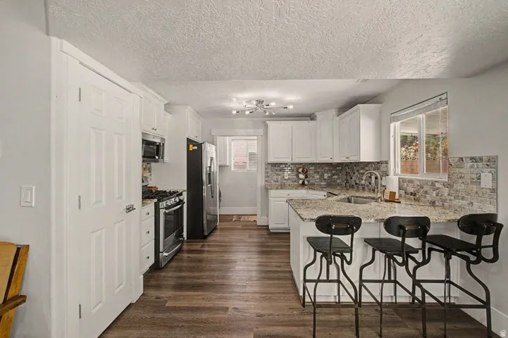 Bright kitchen with white cabinetry, stainless steel appliances, a stone tile backsplash, wood-finish flooring, and a peninsula with seating
