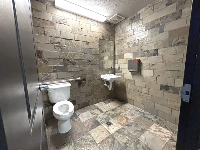Restroom featuring full-height stone wall tiling, coordinating stone floor tiles, a wall-mounted toilet with grab bar, and a wall-mounted sink with mirror