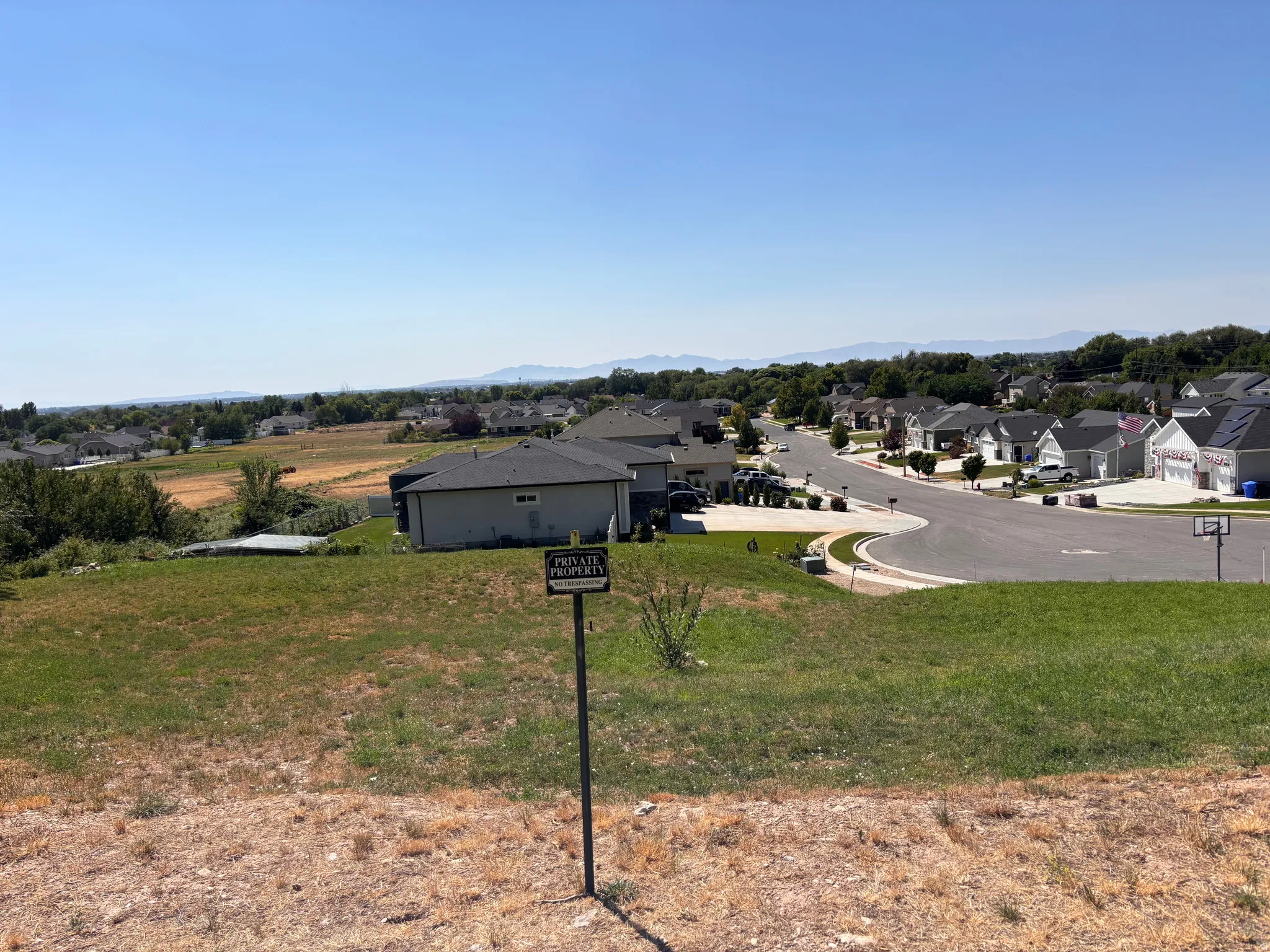 Expansive lot with varied terrain, featuring green and dry grass, a partial residential street, and distant mountain backdrops