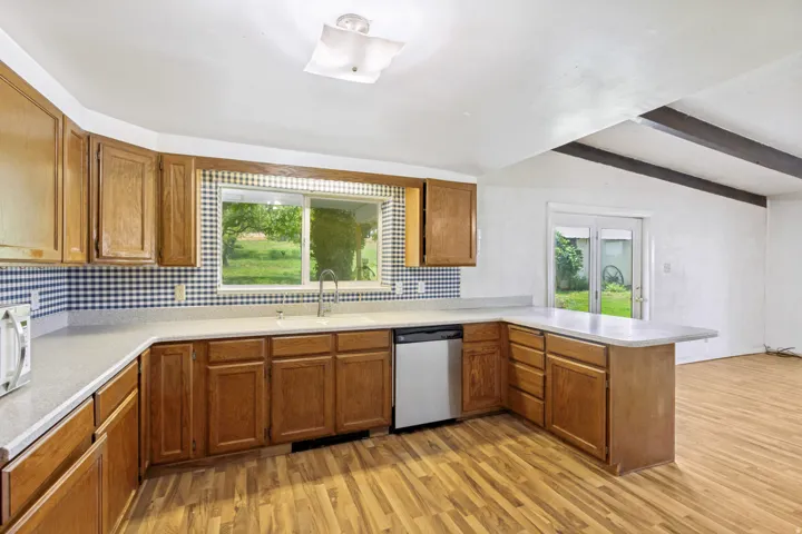 Kitchen featuring a U-shaped countertop configuration, wood-finish flooring, a stainless steel dishwasher, wood cabinetry, and a large window above the sink