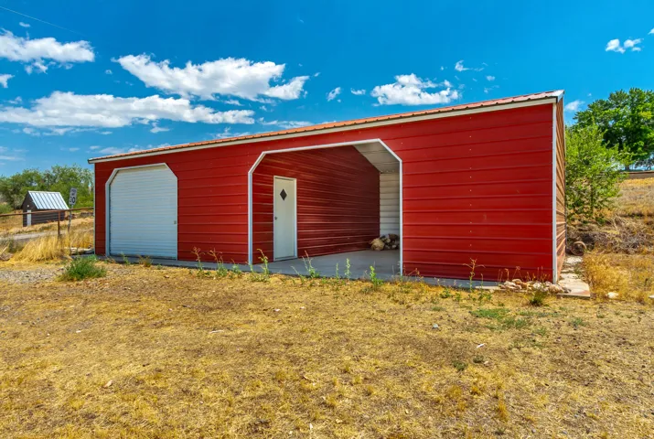 Red metal outbuilding featuring a white roll-up garage door, an open carport, and a white entry door with a diamond-pane window