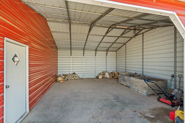 Open-sided metal structure with a concrete floor, featuring a red corrugated metal exterior wall and a white metal entry door with a diamond-shaped window pane