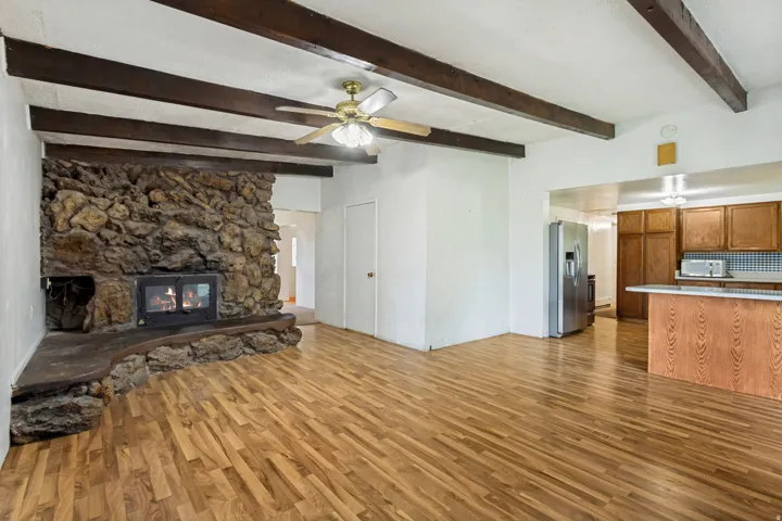 Spacious living area featuring a stone-clad fireplace with hearth, wood-finish flooring, exposed ceiling beams, and a ceiling fan