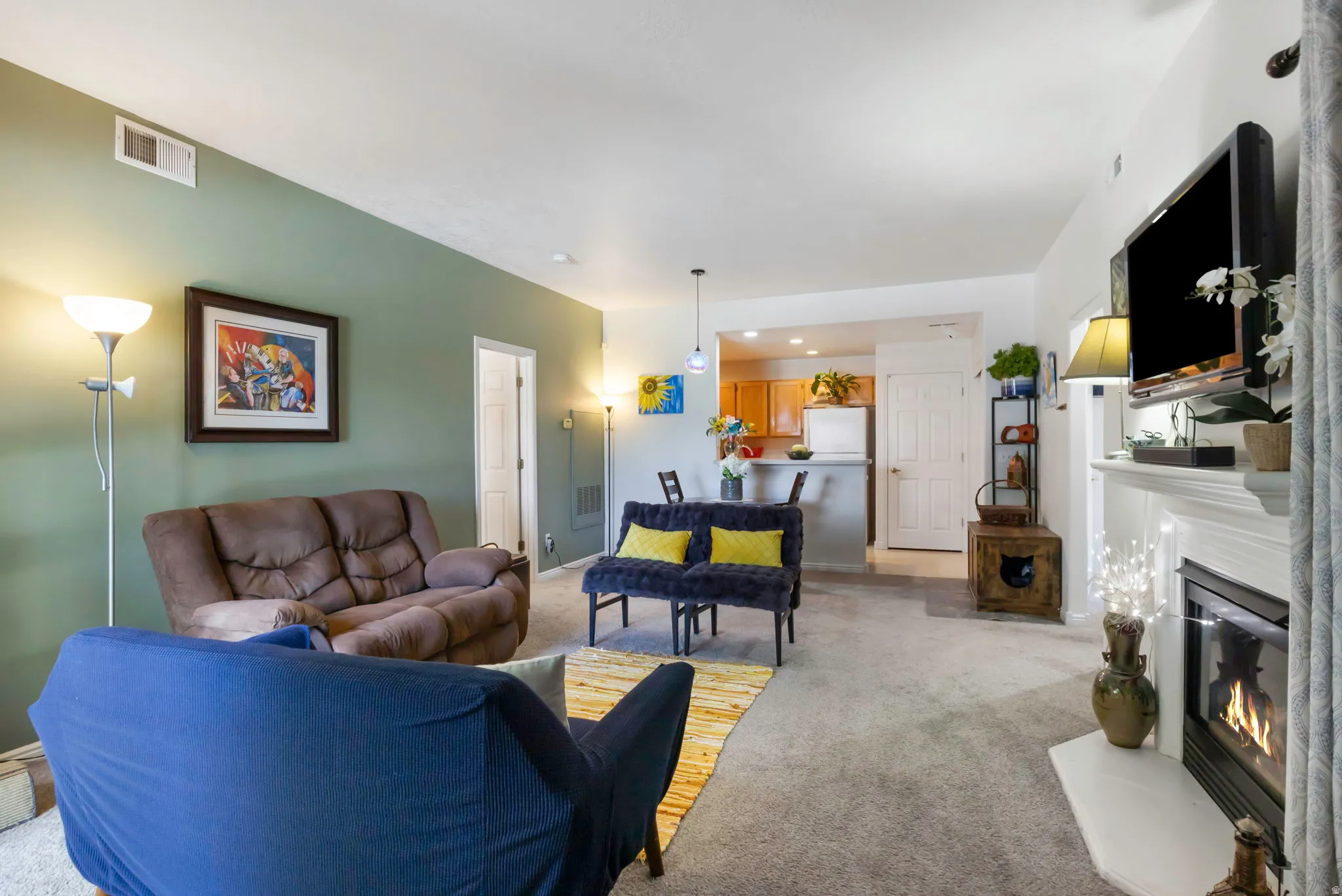 Spacious living area featuring a fireplace with a white mantel, overhead lighting, and light-colored carpeting