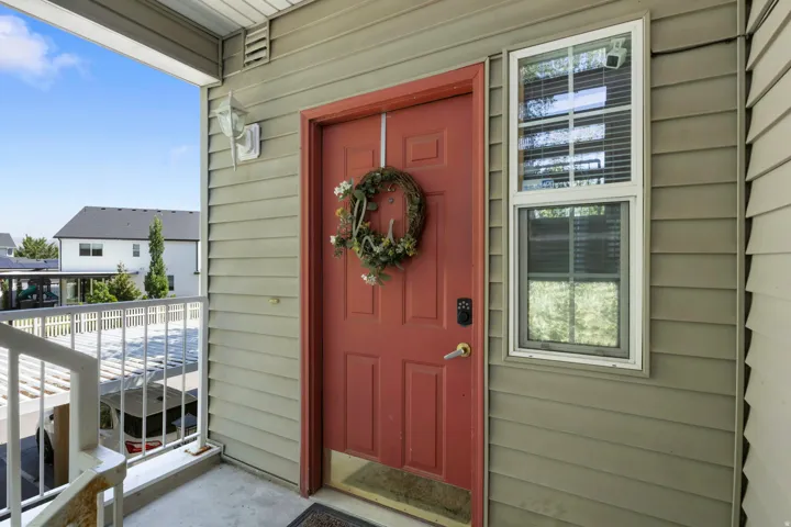 Covered entry with concrete flooring, framed by a red paneled door with smart lock hardware and a white-framed window