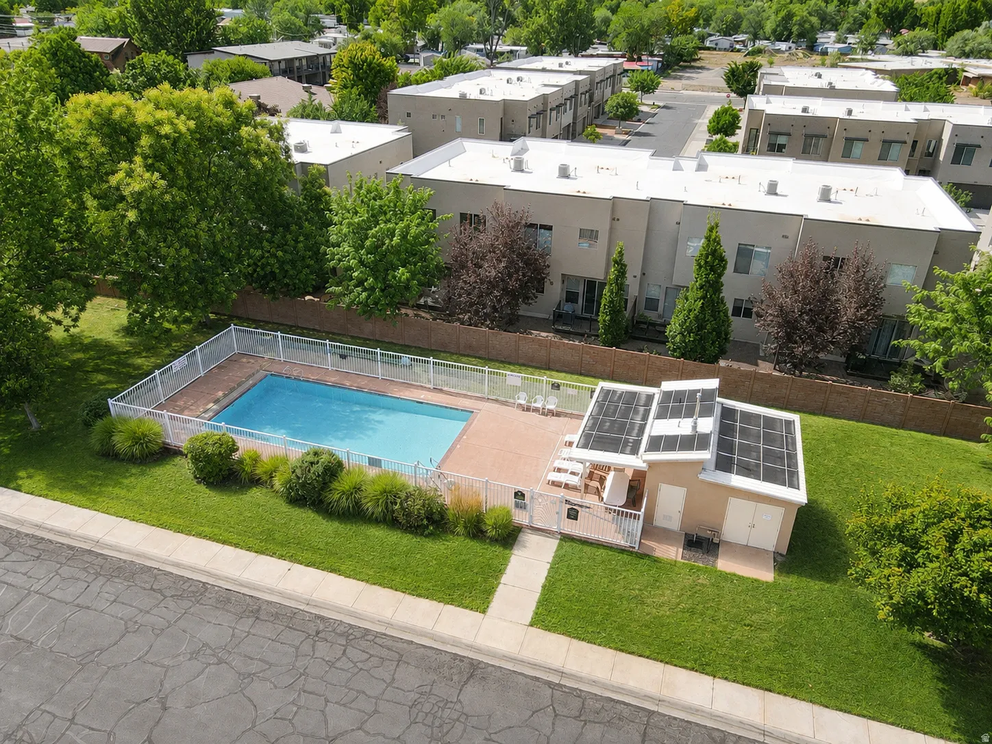 Fenced swimming pool with a surrounding paved deck, adjacent pool house featuring solar panels, and manicured green lawn