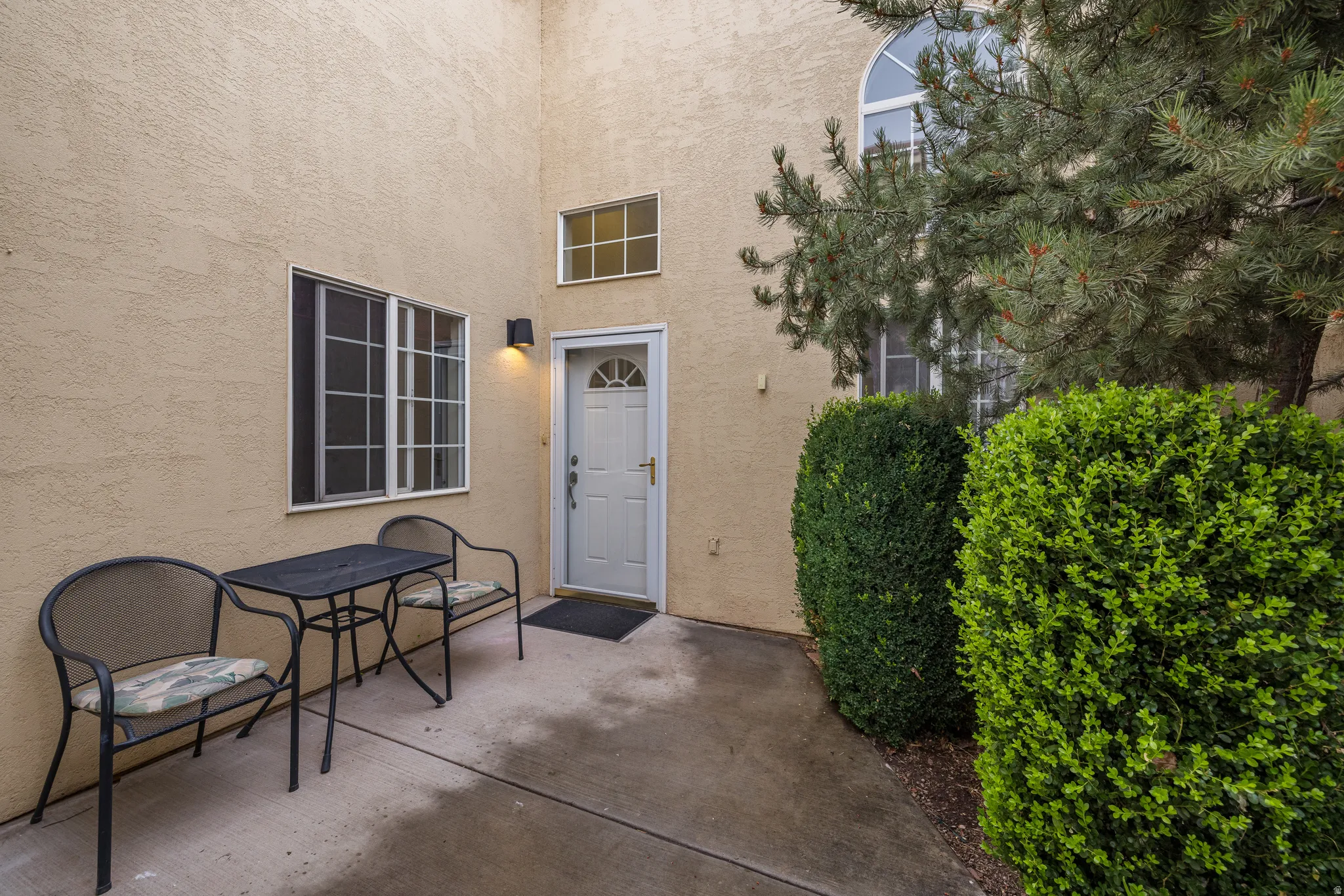 Stucco exterior featuring a white paneled entry door with arched window detail, an exterior wall sconce, and mature perimeter landscaping