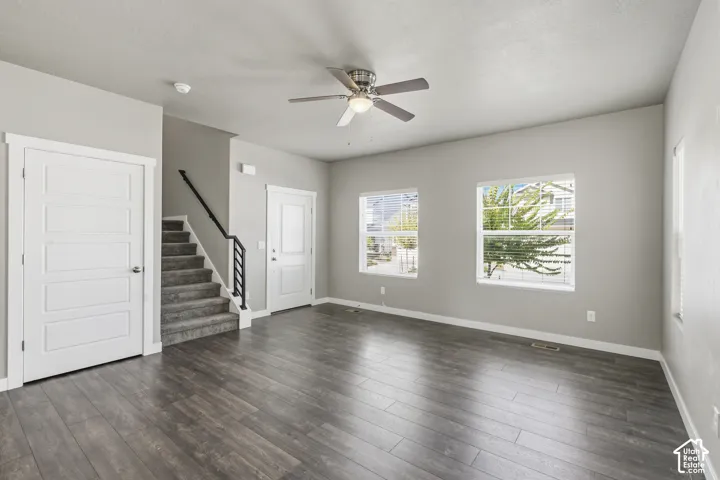 Living area featuring wood-finish flooring, neutral wall paint, two dual-pane windows, a ceiling fan with integrated lighting, and a staircase with a black metal railing
