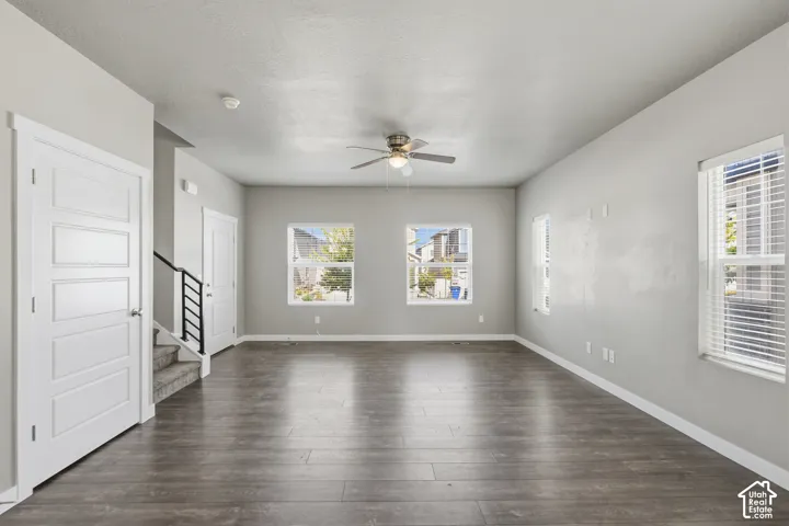 Open-concept interior featuring wood-finish flooring, neutral wall paint, multiple windows with horizontal blinds, a ceiling fan with integrated lighting, and a staircase with a matte black metal railing