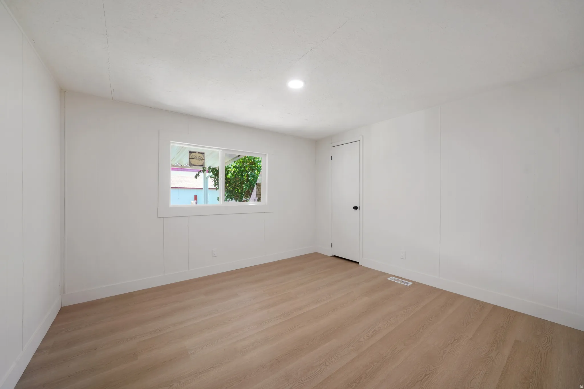 Spacious room featuring light wood-finish flooring, white paneling on walls, a bright window, recessed ceiling lighting, and white baseboards