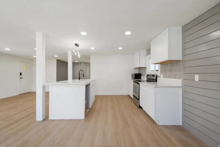 Open-concept kitchen featuring an island with a built-in sink, white cabinetry, stainless steel appliances, a light-colored wood-finish floor, and a gray shiplap accent wall