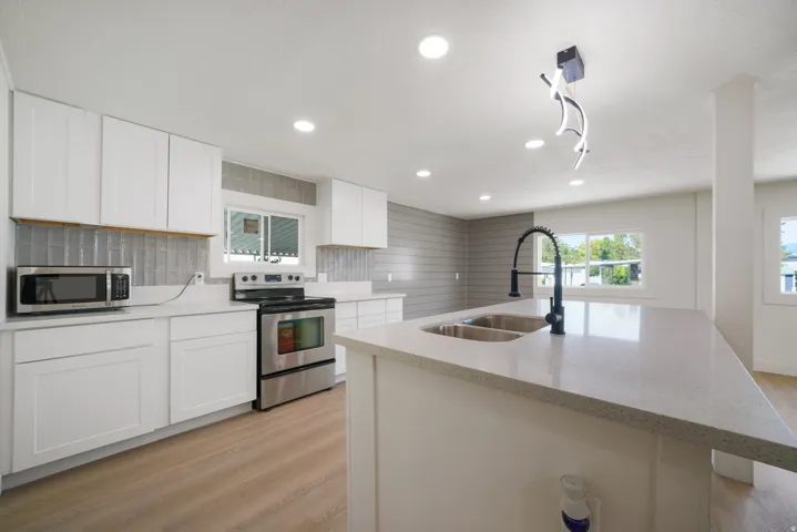 Open-concept kitchen featuring an island with a double basin sink and gooseneck faucet, light-toned wood-finish flooring, white cabinetry, stainless steel appliances, and recessed lighting