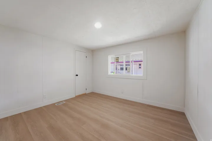 Bright room featuring light wood-finish flooring, white paneled walls, a white door with black hardware, and a window with white trim