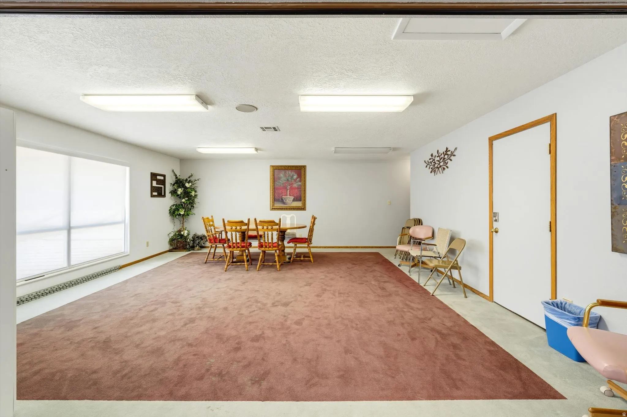 Spacious room featuring a wide window with roller shades, white walls, and a large area rug