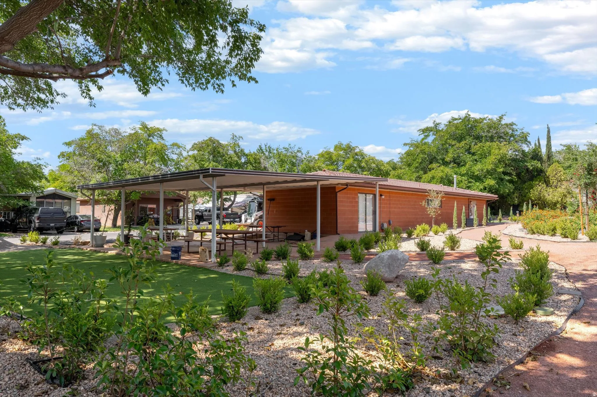 Wood-finish exterior structure with a covered patio area, artificial turf lawn, landscaped rock garden, and mature trees