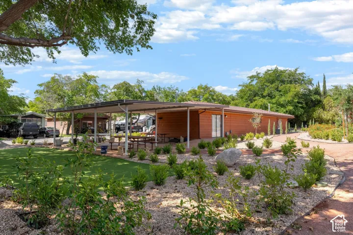 Wood-finish exterior structure with a covered patio area, artificial turf lawn, landscaped rock garden, and mature trees