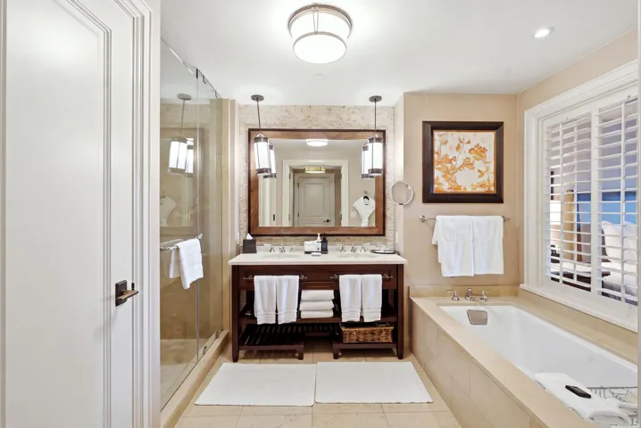 Full bathroom featuring a single vanity with stone countertop, frameless glass shower enclosure, built-in bathtub, and window with plantation shutters