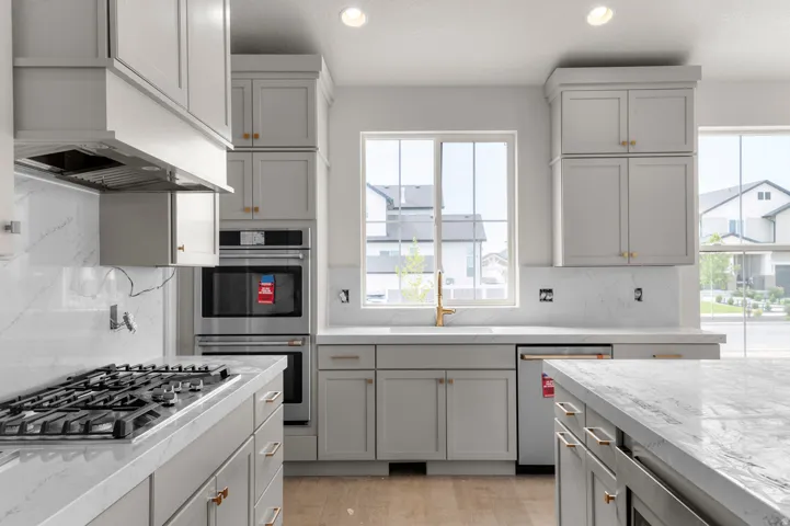 Modern kitchen featuring light gray cabinetry with gold-toned hardware, quartz countertops, a gas range, a built-in double oven, and a large window above the sink