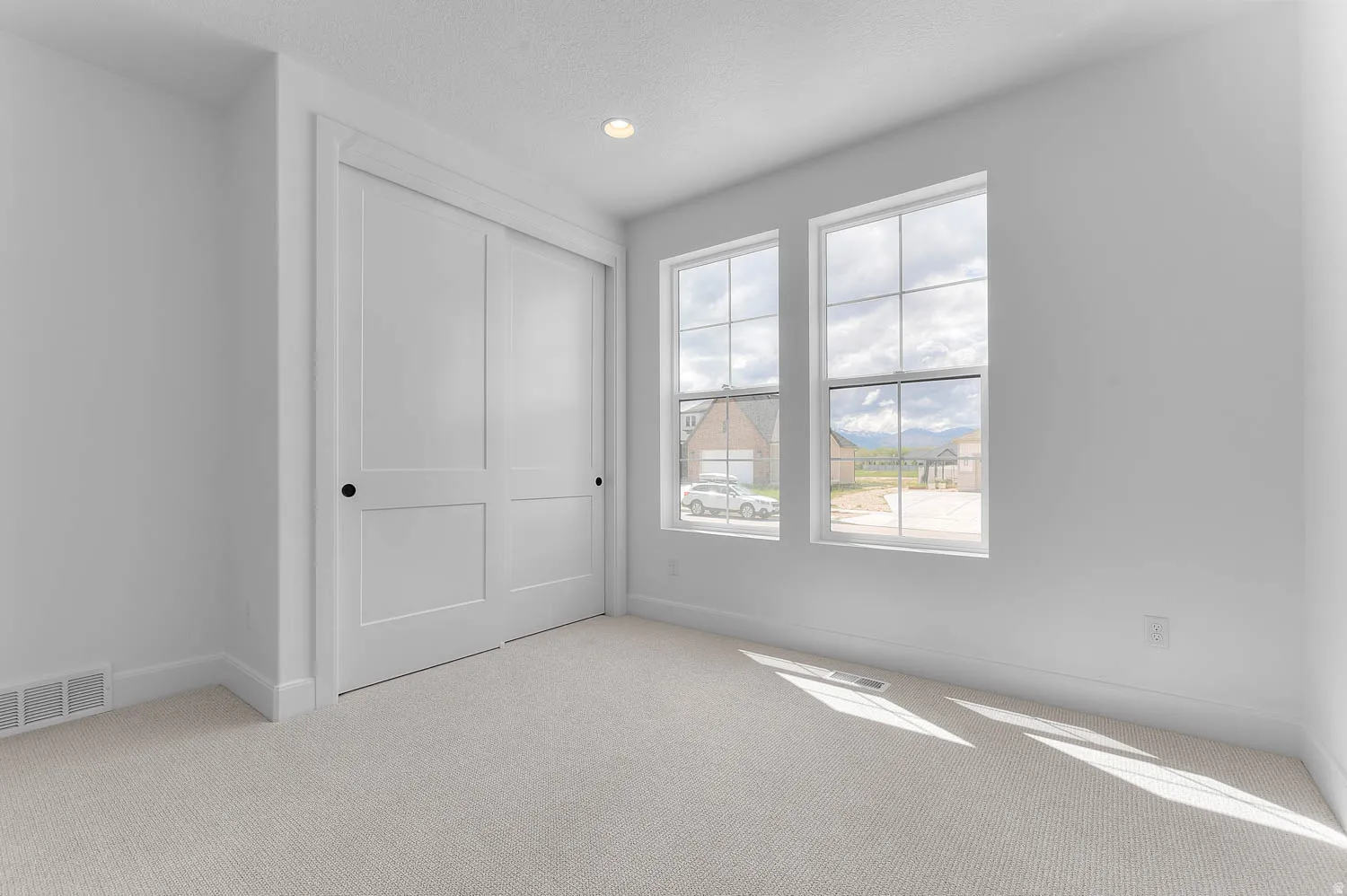 Recessed lighting illuminates a space featuring two large windows, a sliding two-panel closet door, and light-toned carpeting