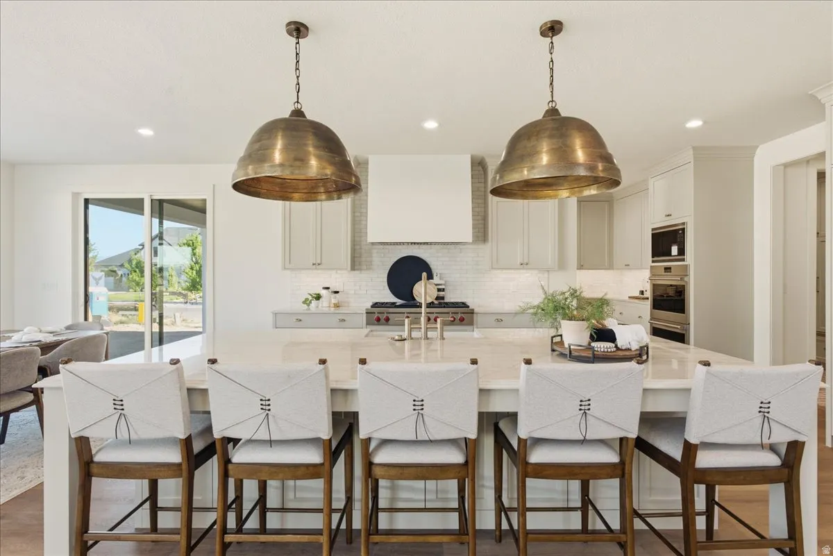 Kitchen featuring a large central island with seating, pendant lighting fixtures, light-toned cabinetry, a built-in range hood, and a tile backsplash