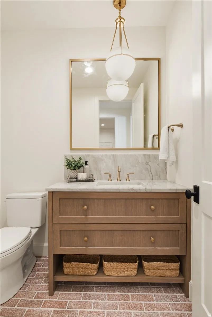 Powder room featuring brick flooring, a wood-finish vanity with a stone countertop and backsplash, a rectangular mirror with a gold-tone frame, and a gold-tone pendant light fixture