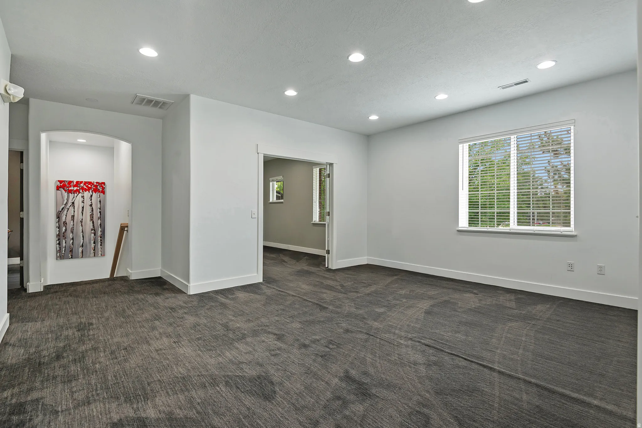 Spacious room featuring gray patterned carpeting, recessed lighting, white trim, a large window with blinds, and an arched wall niche