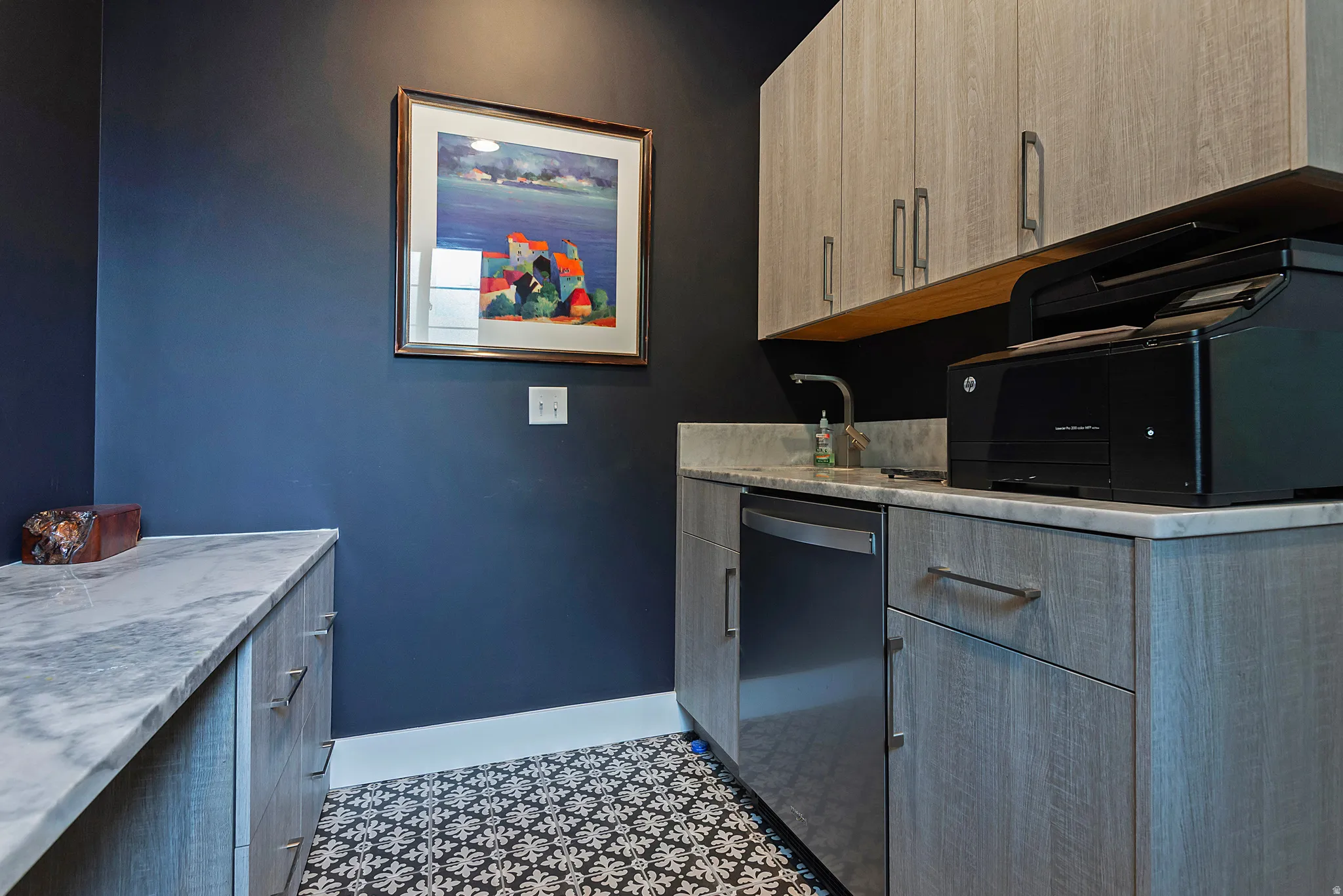 Utility area featuring patterned floor tile, blue accent wall, light wood-finish cabinetry with silver hardware, and stone countertops