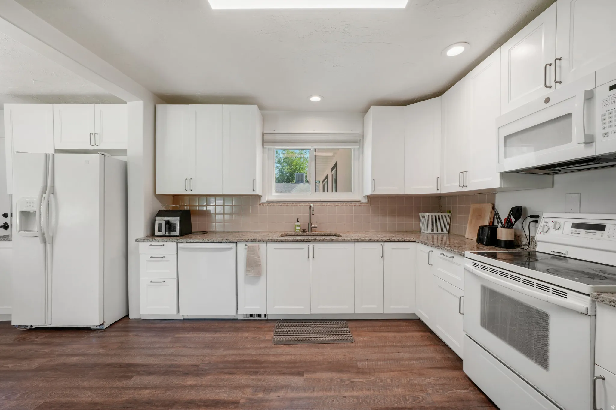 Kitchen featuring wood-finish flooring, white cabinetry, speckled countertops, a tile backsplash, and a window above the sink