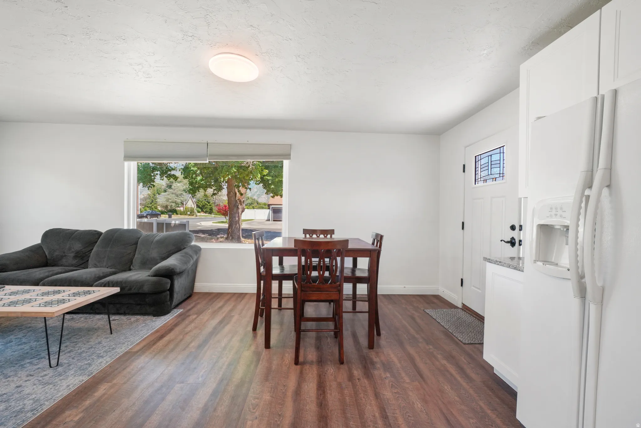 Open-concept living space featuring wood-finish flooring, white walls, and a large front-facing window with roller shades