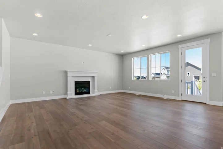 Spacious living area featuring light wood-finish flooring, a contemporary fireplace with a classic white mantel, recessed lighting, and multiple large windows for abundant natural light