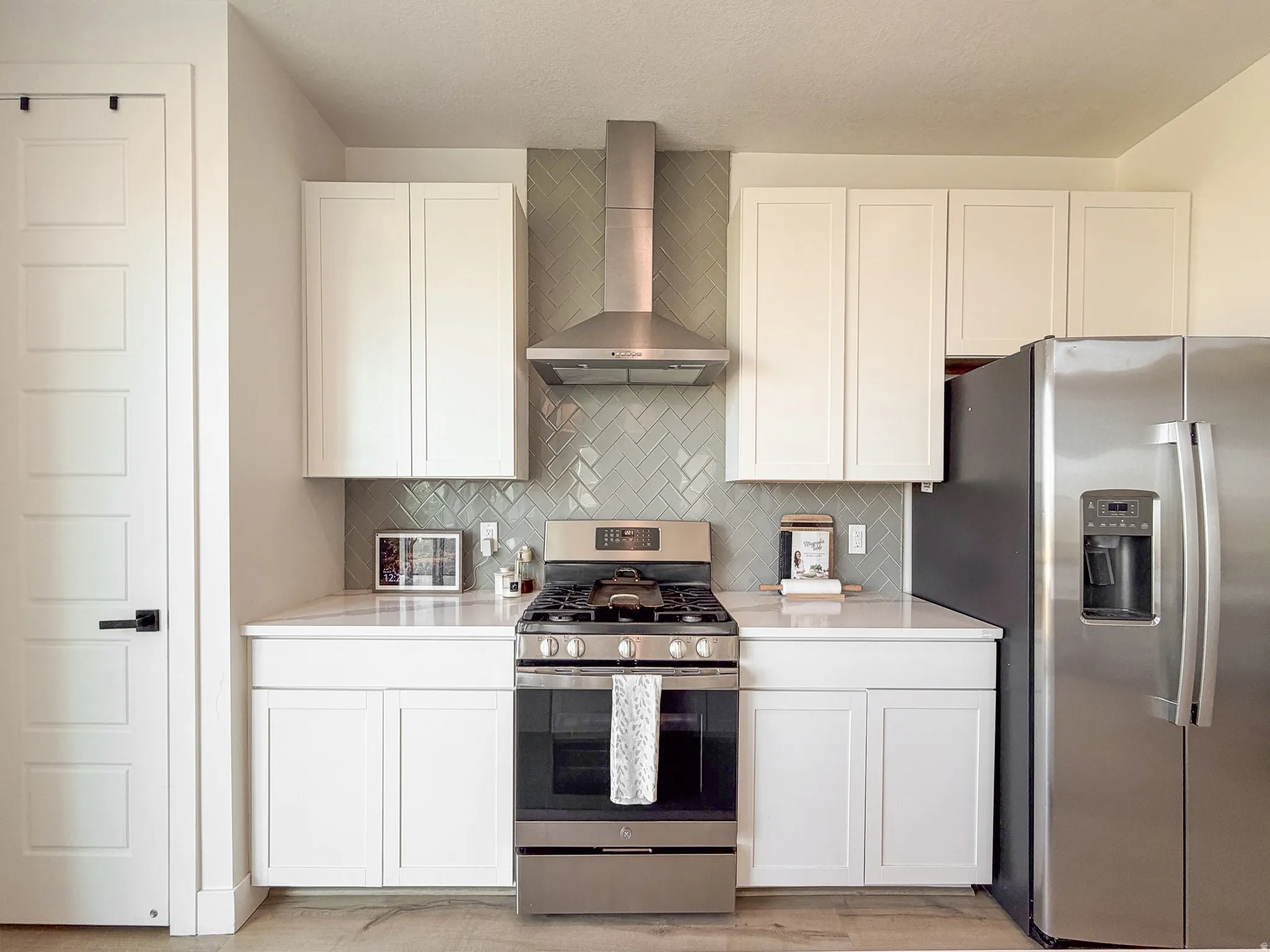 Modern kitchen featuring white shaker cabinetry, stainless steel appliances, a range hood, and a light gray herringbone tile backsplash