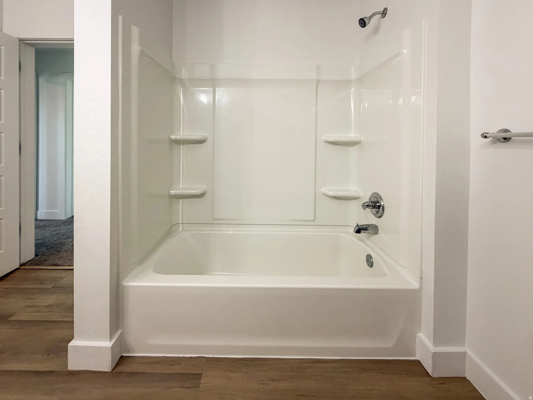 White bath and shower combination with chrome fixtures and integrated shelving, set against wood-finish flooring