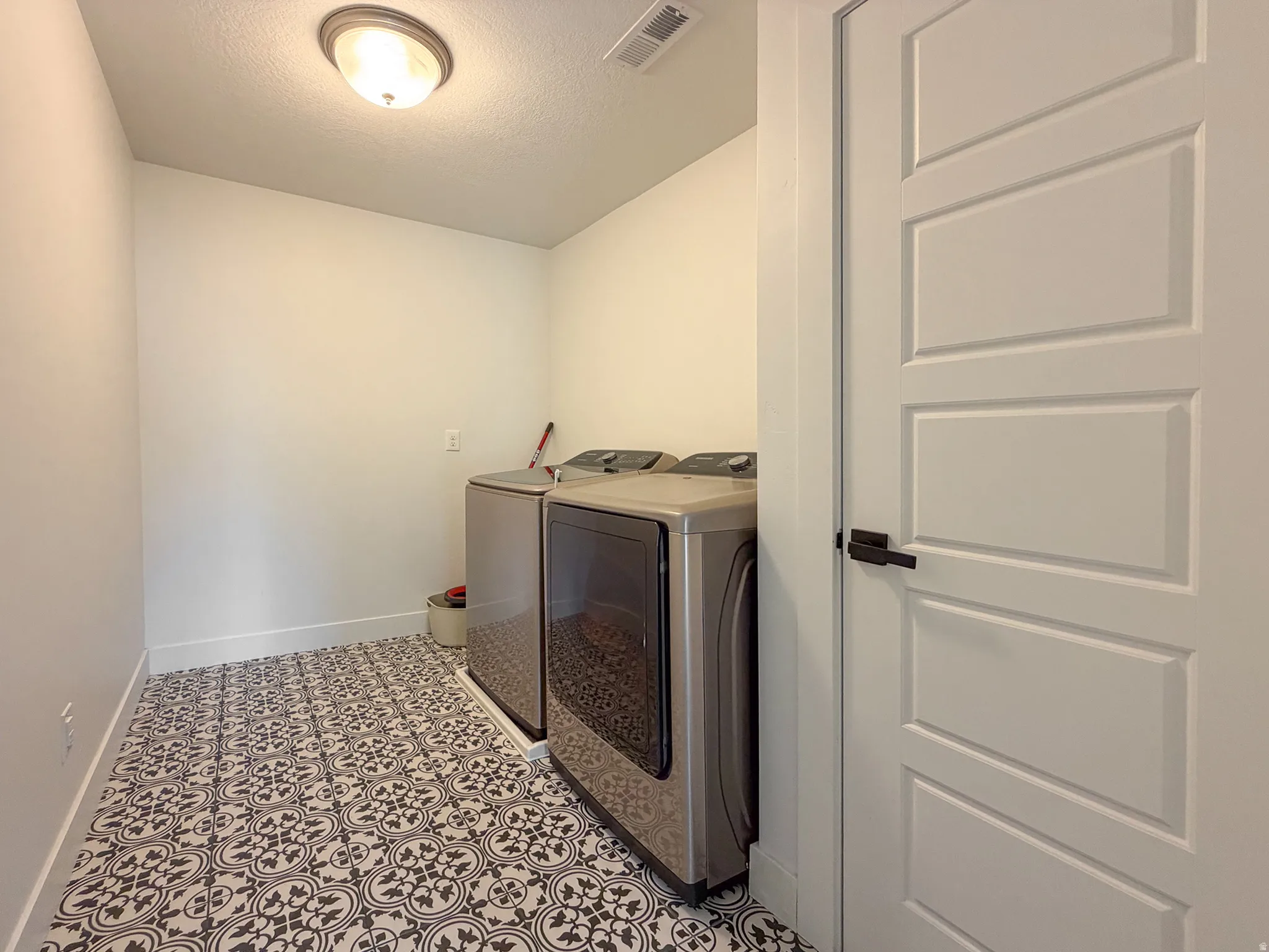 Utility room featuring patterned tile flooring, a flush-mount ceiling light fixture, white baseboards, and a paneled interior door with matte black hardware